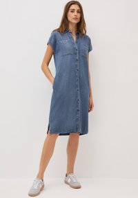 Abito camicia in denim, azzurro chiaro, maniche corte, chiusura con bottoni, due tasche sul petto, fessure laterali, abbinato a sneaker grigie. Texture liscia.