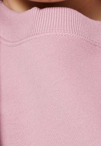 Ljusrosa tröja med en mjuk textur, som har en ribbad halsringning och subtila sömdetaljer. Tyget verkar slätt och andningsbart.