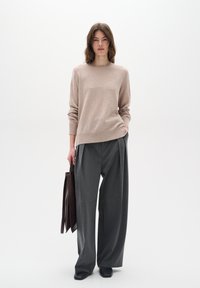 Pull en tricot beige à manches longues, associé à un pantalon gris ample. Le modèle tient un sac fourre-tout marron foncé ; arrière-plan minimaliste.