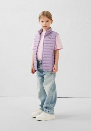 Kind draagt lichtpaars gewatteerd vest over roze T-shirt, losse lichtblauwe spijkerbroek, witte sneakers, staand tegen een effen witte achtergrond.
