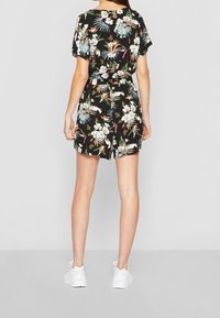Mujer vestida con un mono corto de manga corta negro con flores, cinturón y zapatillas blancas, de pie frente a un fondo gris claro liso.