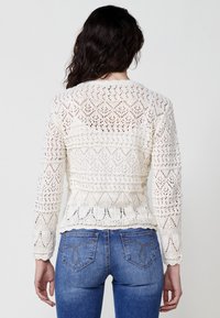 Suéter blanco de punto con un diseño de ganchillo que presenta patrones geométricos y mangas largas, usado sobre unos jeans azules.