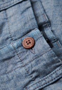 Denim w jasnoniebieskim kolorze z brązowym guzikiem z wygrawerowanym napisem "Superdry". Zbliżenie na szczegóły przeszyć w pobliżu guzika.