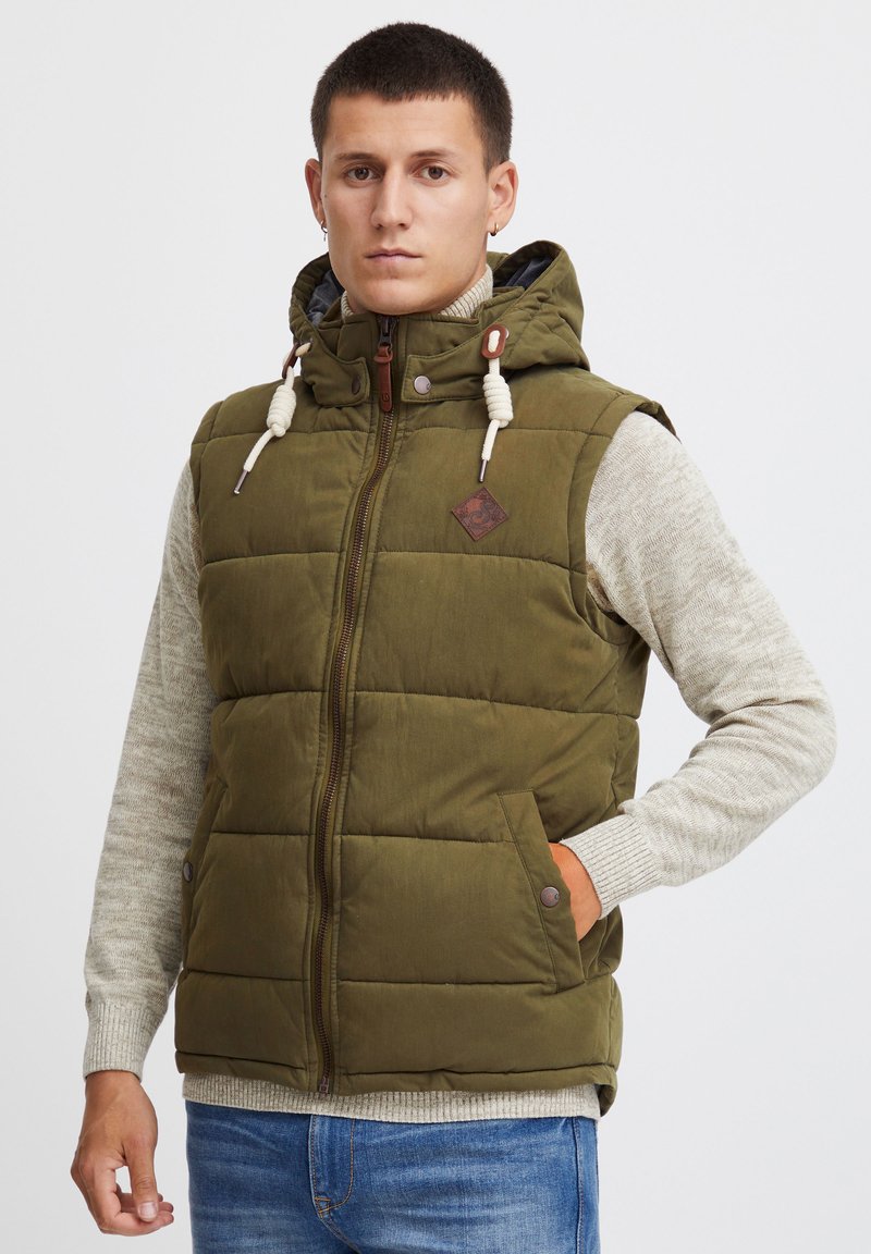 Gilet matelassé vert olive avec capuche, doté d'une fermeture éclair, de deux poches latérales et de détails en cuir marron. Porté par-dessus un pull léger.