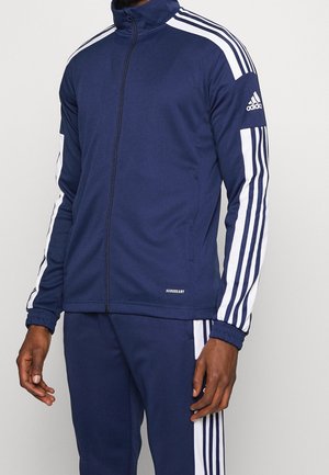 Dunkelblauer Adidas-Trainingsanzug mit weißen Streifen an Ärmeln und Hosen, durchgehendem Reißverschluss und elastischen Bündchen, getragen von einer Person mit dunkler Haut.