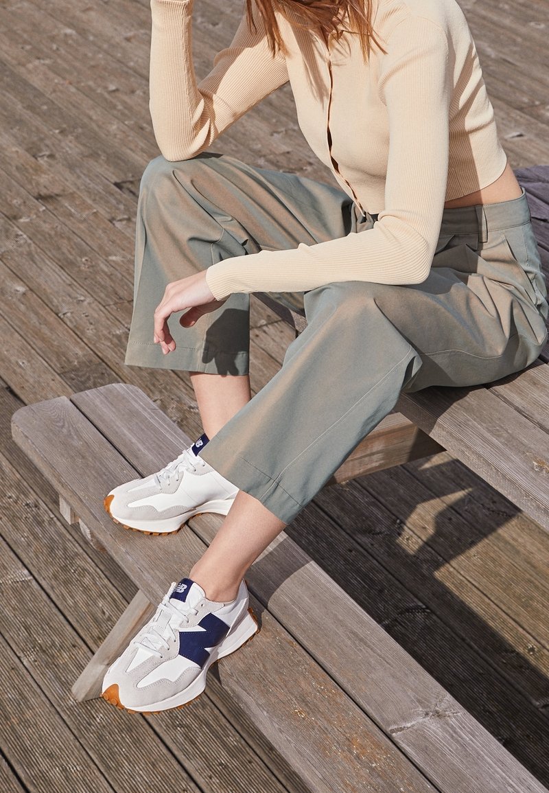 Zapatillas blancas y grises con detalles en azul marino y suela naranja, combinadas con pantalones anchos de pierna ancha en verde claro y una camiseta de canalé beige, sentada sobre tablones de madera.