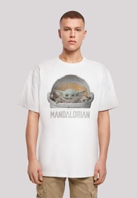 Camiseta blanca de algodón con un gráfico de un personaje verde en una cápsula, con el texto "EL MANDALORIANO" debajo, sobre un fondo neutro.