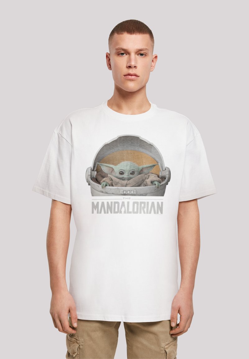 Camiseta blanca de algodón con un gráfico de un personaje verde en una cápsula, con el texto "EL MANDALORIANO" debajo, sobre un fondo neutro.