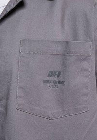Grå denim arbejdsskjorte med spidse krave, brystlomme og trykt logo "DEF WORKATION MODE F/W23" på lommen.
