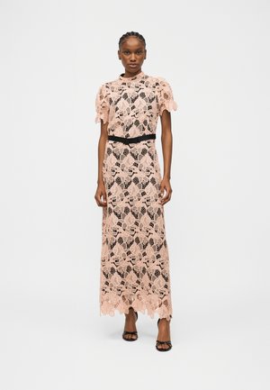 Kvinde iført gulvlange lyserøde blondekjole med pufærmer og sort bælte, stående med neutral udtryk og sorte åbne hæle.