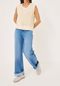 Maglione senza maniche beige lavorato a maglia con scollatura a V, abbinato a jeans a gamba larga azzurri chiari e sneakers grigie con un design discreto.