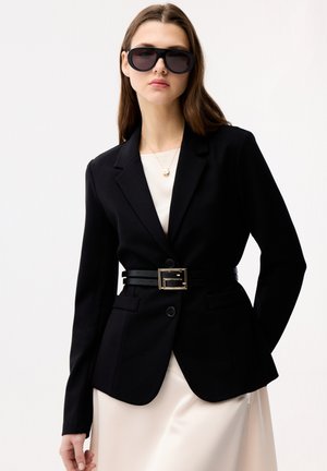 Woman wearing black tailored blazer with double-belted waist, cream dress, black oversized sunglasses, and a gold pendant necklace.