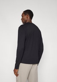 Maglietta nera a maniche lunghe con scollo rotondo, caratterizzata da una texture liscia e una vestibilità comoda. Indossata con pantaloni grigio chiaro.