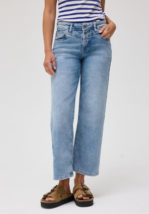 Person iført lyseblå vidde jeans, stribet skjorte og brune plateau-sandaler med spænder, der står med den ene hånd i lommen.