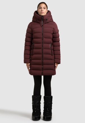 Žena v dlouhém vínovém zatepleném zimním kabátu s vysokým límcem, černých legínách a černých sněhobotách na šněrování, stojící proti šedému pozadí.