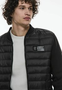 Svart pufferjacka med en texturerad sidopanel, dragkedja fram och ett märkespatch på vänster bröst. Bärs över en vit texturerad skjorta.