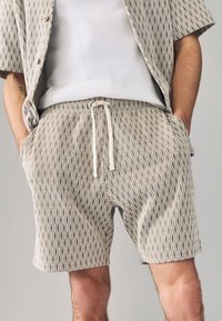 Beige korte broek met een diamantpatroon, voorzien van een elastische tailleband en trekkoord. Gecombineerd met een wit hemd eronder.