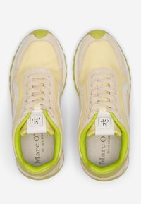 Beige sneakers with yellow accents and white side stripes. Rounded shape, textured fabric, and a prominent green inner lining.