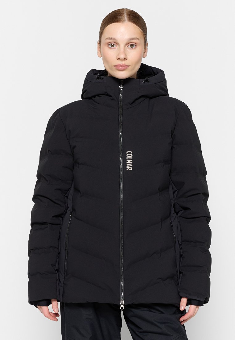 Giovane donna in un giubbotto imbottito nero con cappuccio, chiusura frontale con zip e logo "COLMAR" verticale, in piedi davanti a uno sfondo bianco semplice.