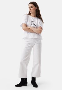 T-shirt bianco cropped con stampa grafica nera, abbinato a pantaloni bianchi a gamba larga e stivali neri alla caviglia. Texture del tessuto lisce.