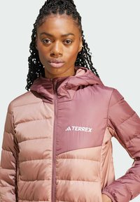 Giacca imbottita nelle tonalità di rosa con chiusura a zip e cappuccio. Presenta un logo sul lato sinistro del petto e una texture trapuntata.