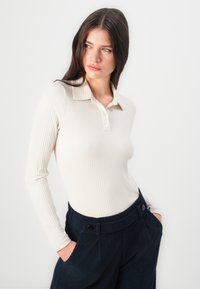 Polo a coste di colore crema con colletto corto e patta con bottoni anteriore, abbinato a pantaloni in velluto a coste di colore blu scuro con tasche laterali e bottoni.