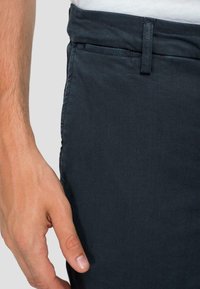 Pantaloni in cotone blu scuro con una consistenza liscia. Presentano una vita standard e tasche laterali. Mano posizionata vicino al tessuto.