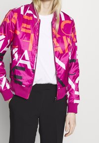Felroze bomberjack met opvallende zwarte en oranje grafische tekst. Voorzien van een ritssluiting, geribbelde manchetten en een gladde, glanzende stof.
