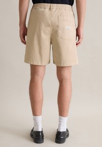 Beige cotton shorts with an elastic waistband, back pocket, and cuffed hem, worn with black shoes and white socks. Model stands facing away.