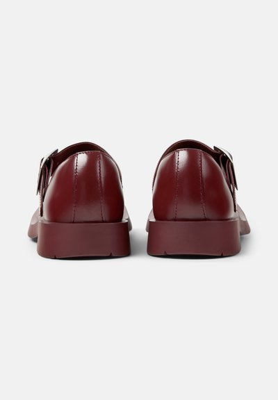 Paire de chaussures en cuir rouge foncé poli avec semelles épaisses et boucles latérales, vues de dos sur un fond blanc.