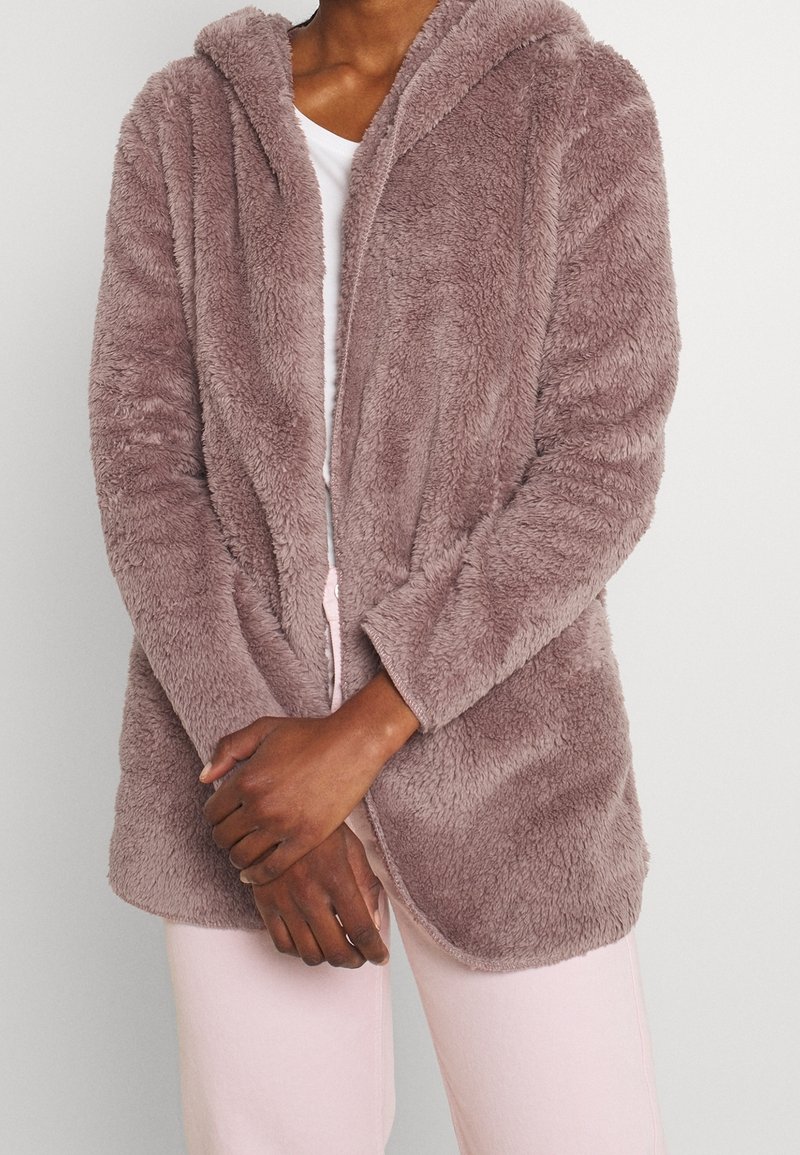 Personne portant une veste à capuche mauve et duveteuse par-dessus une chemise blanche et un pantalon rose clair, debout les mains jointes devant.