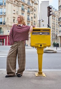 Pasiasty burgundowy top o oversize'owym kroju, zestawiony z szerokimi spodniami w kratę w kolorach brązu i zieleni, stoi obok żółtej skrzynki pocztowej.