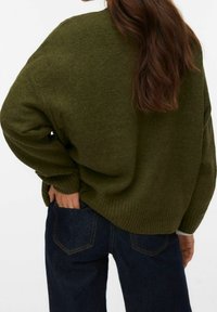 Femme aux longs cheveux bruns portant un pull oversize vert olive et un jean bleu foncé, main dans la poche arrière, vue de dos.
