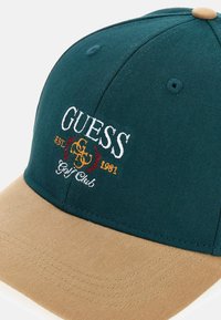Cappello verde scuro con visiera beige, con logo ricamato "GUESS' Golf Club" e dettagli in oro e rosso sul davanti.