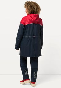 Veste imperméable navy avec une partie supérieure rouge, capuche à cordon de serrage et accents réfléchissants. Associée à un pantalon navy avec des motifs en chevron sur les poignets.