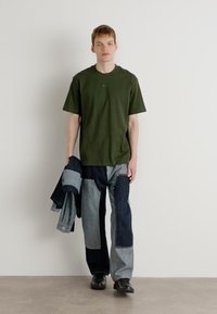 Grünes Baumwoll-T-Shirt, lockere Passform, kombiniert mit Jeanshose im Patchwork-Stil in Blautönen, hält eine blaue Jeansjacke, schwarze Schuhe.