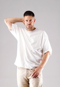 White, short-sleeve t-shirt with a round neckline and subtle logo detail, paired with light beige pants, set against a plain background.