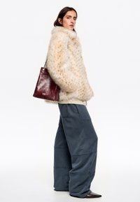 Kurtka z sztucznego futra w kremowym kolorze z brązowymi plamkami, zestawiona z ciemnoniebieskimi spodniami o szerokich nogawkach oraz bordową skórzaną torbą, prezentująca warstwową fakturę.