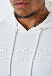 Sudadera blanca con un logo en relieve "PROJECT X". Incluye cordones y una textura suave, con un primer plano del cuello y tatuajes visibles.