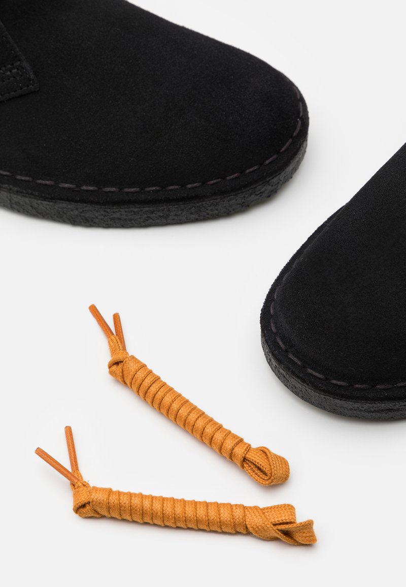 Zwarte suède schoenen met rubberen zolen, voorzien van dunne oranje veters met gedraaide uiteinden. Lage snit met minimale stikdetails.