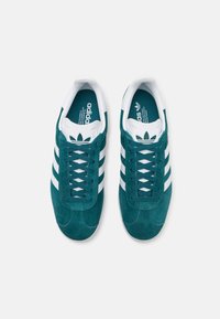 Gröna mockasneakers med tre vita ränder, snörning och vadderad plös. Har en blå innerdel med logotyp och texturerad gummisula.