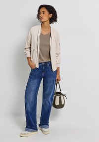 Veste bomber beige clair avec fermeture éclair sur le devant, manches courtes ; top taupe à col en V ; jeans larges bleus ; baskets blanches ; petit sac à main vert.