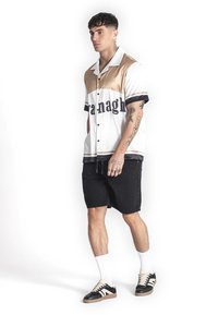 Camisa de manga corta con botones en blanco, dorado y negro con un gran diseño de texto; combinada con pantalones cortos negros y zapatillas negras.