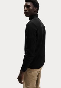 Suéter negro de punto con cuello alto y mangas largas, con un cuello y dobladillo acanalados. Llevado con pantalones de color claro. Diseño simple y moderno.