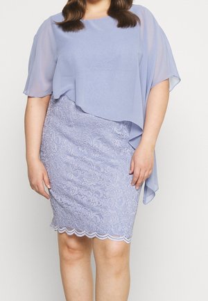 Kvinde iført en lyse lilla blondekjole med en gennemsigtig, asymmetrisk overlay-top, stående mod en ensfarvet lys baggrund.