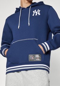 Felpa con cappuccio blu navy con logo bianco, cordini e polsi a righe. Presenta una tasca frontale e una patch con il marchio sull'orlo. Tessuto morbido.