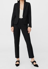 Blazer noir ajusté avec un seul bouton, associé à un pantalon noir ajusté. Haut léger texturé en dessous ; talons noirs pointus avec des brides à la cheville.