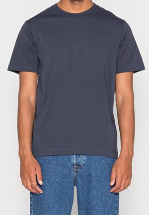 Mand iført en ensfarvet mørkegrå kortærmet t-shirt med rund hals og blå denimjeans, armene afslappet ned langs siderne.