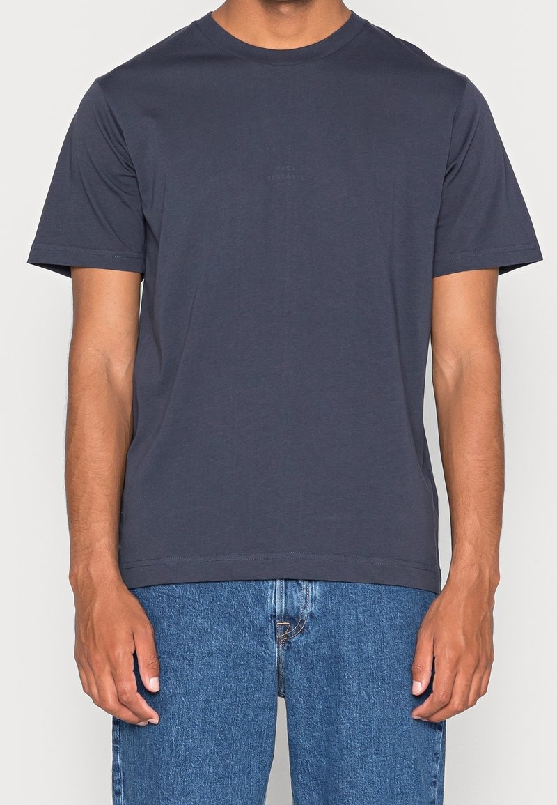 Homme portant un t-shirt gris foncé uni à manches courtes et col rond, et un jean en denim bleu, bras détendus sur les côtés.