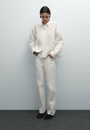 Chemise en coton blanc avec un devant texturé, coupe décontractée ; associée à un pantalon en denim blanc et des chaussures noires à enfiler. Fond neutre.